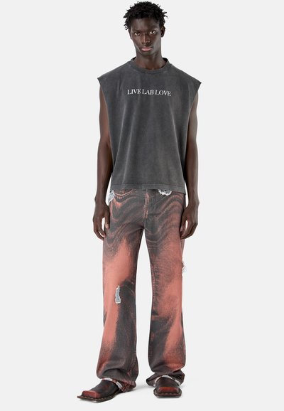 Jonge man draagt een zwarte mouwloze T-shirt met de tekst "LIVE LAB LOVE", versleten wijde jeans met een oranje-zwart patroon en zwarte schoenen.