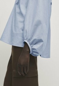 Personne à la peau foncée portant un chemisier bleu clair à manches longues avec des poignets froncés et un pantalon marron foncé avec un rabat de poche.