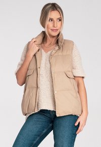 Beżowa kamizelka puchowa z wysokim kołnierzem, zatrzaskami i dwiema przednimi kieszeniami, nałożona na lekką kremową sweter z dzianiny i niebieskie dżinsy.