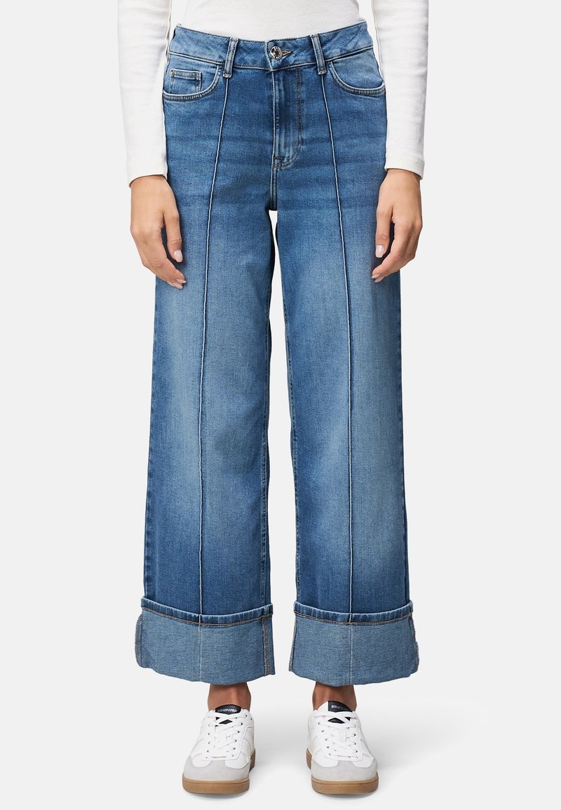 Femme portant un jean bleu taille haute à jambes larges avec coutures frontales et grands revers, associé à des baskets blanches et un haut blanc à manches longues.
