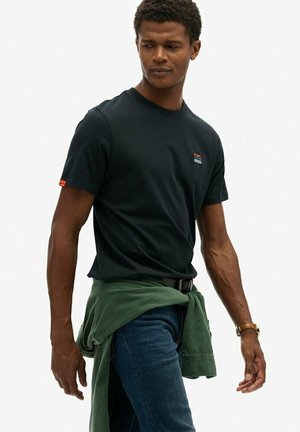 T-shirt en coton noir à manches courtes, avec un petit logo multicolore sur la poitrine et un tissu vert roulé noué autour de la taille.