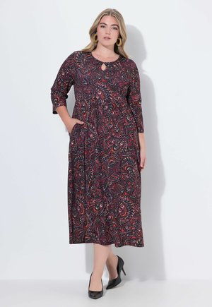 Robe noire avec un motif paisley coloré, manches 3/4, décolleté en forme de trou de serrure, poches latérales et une coupe fluide. Portée avec des talons hauts noirs.