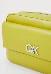 Borsa in pelle verde lime con una texture liscia, dotata di un logo argentato e chiusura a zip. Il design è rettangolare con un cappuccio ripiegabile.