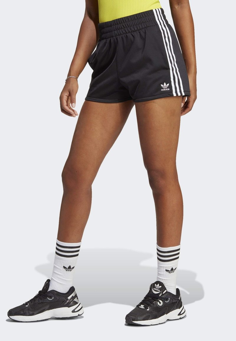 Ben iklädda svarta Adidas-shorts med vita ränder, vita Adidas-strumpor med svarta ränder och svarta Adidas-sneakers mot en ljus bakgrund.