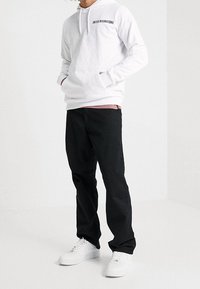 Biała bluza z kapturem z napisem "UNITE INTERNATIONAL", zestawiona z czarnymi spodniami i białymi trampkami, stojąca na jasnym tle.