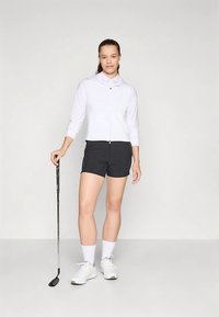Maglia bianca con zip e maniche lunghe abbinata a pantaloncini neri, che tiene una mazza da golf. Le scarpe sono bianche con dettagli grigi e indossa calzini bianchi.