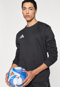 Μαύρο φούτερ με μακριά μανίκια, στρογγυλή λαιμόκοψη και αντανακλαστικό λογότυπο Adidas. Άτομο κρατά μια πολύχρωμη ποδοσφαιρική μπάλα με στροβιλισμένα σχέδια.