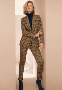 Bruin gestreept pak met een getailleerde blazer en nauwsluitende broek, aangevuld met een donkere coltrui en marineblauwe hakken. Textuur van de stof is zichtbaar.