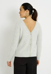 Cárdigan gris claro acanalado con un escote profundo en V en la espalda, que presenta un cierre de botones y un detalle dorado a lo largo de la columna vertebral. Tejido suave y texturizado.