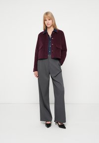 Giacca di velluto a coste bordeaux con due tasche frontali, bottoni argento, abbinata a pantaloni grigi a gamba larga e scarpe nere con tacco.