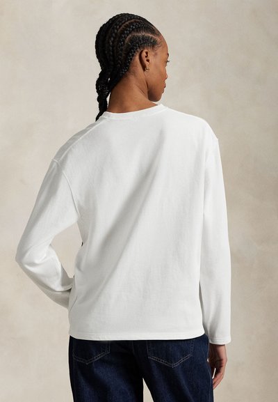 Kobieta z warkoczami stoi tyłem, ubrana w prostą białą bluzkę z długim rękawem i ciemnoniebieskie dżinsy na neutralnym tle.