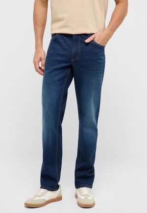 Mann trägt blaue Jeans mit geradem Bein, beige T-Shirt und weiße Turnschuhe mit beigen Akzenten, steht mit einer Hand in der Tasche.