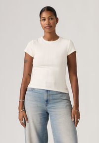 Γυναίκα με σκούρα μαλλιά, φορώντας λευκό τοπ με κοντά μανίκια και ανοιχτόχρωμο μπλε τζιν, όρθια με τα χέρια χαλαρά δίπλα στο σώμα της, μπροστά σε μονόχρωμο φόντο.