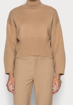 Person wearing a camel-colored turtleneck sweater and matching pants with a gold bracelet and ring on their right hand.