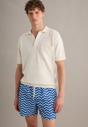 Uomo che indossa una polo bianca a maniche corte in maglia con taschino sul petto e pantaloncini da bagno blu con motivi ondulati bianchi e coulisse.