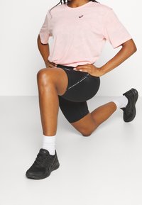 Pink athletic t-shirt with a subtle pattern, paired with black bike shorts featuring a white logo. Black sports shoes and white socks.
