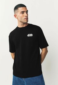 Tricou din bumbac negru cu mâneci scurte, având un logo alb "STAR WARS" pe partea stângă a pieptului, cu o croială lejeră și guler rotund.