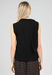 Pull en tricot sans manches noir avec une texture douce, présentant un long dos drapé et porté sur une jupe à pois.