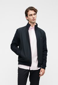 Νεαρός άνδρας με σγουρά μαλλιά, φορώντας μαύρο μπουφάν με φερμουάρ πάνω από ροζ κουμπωτό πουκάμισο και μαύρα παντελόνια, στέκεται με το ένα χέρι στην τσέπη.