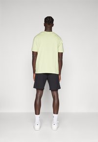 Light green short-sleeve t-shirt paired with black shorts, visible back view. Model wearing white sneakers and socks. Simple texture, relaxed fit.
