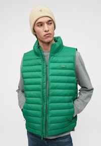 Vestă verde cu umplutură și cusături orizontale, guler înalt și închidere cu fermoar. Asortată cu o bluză gri cu mânecă lungă și o căciulă tricotată bej.