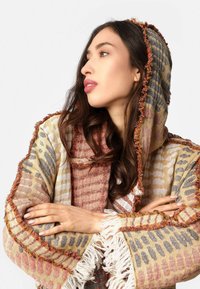 Tkaný šál s kombináciou vzorov v béžovej, sivej a hrdzavohnedej farbe. S franjami na okrajoch a kapucňou. Mäkko textúrovaný a vrstvený dizajn.