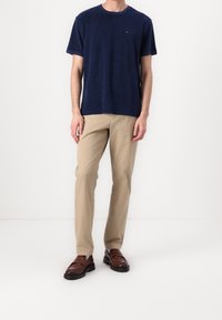 Mann in hellblauem kurzärmligen Hemd, beige geraden Hosen und braunen Lederloafers, der vor weißem Hintergrund steht.