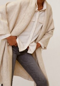 Personne portant un cardigan en tricot beige sur une chemise blanche à boutons et un jean gris, les mains dans les poches, se tenant contre un fond neutre.
