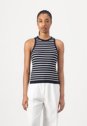 Vrouw staat en draagt een mouwloos marineblauw-wit gestreept gebreid topje, gecombineerd met witte broek met hoge taille tegen een effen achtergrond.