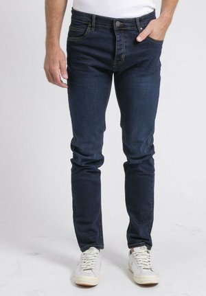 Homme portant un jean slim bleu foncé et des baskets blanches, debout avec une main dans la poche devant un fond uni.