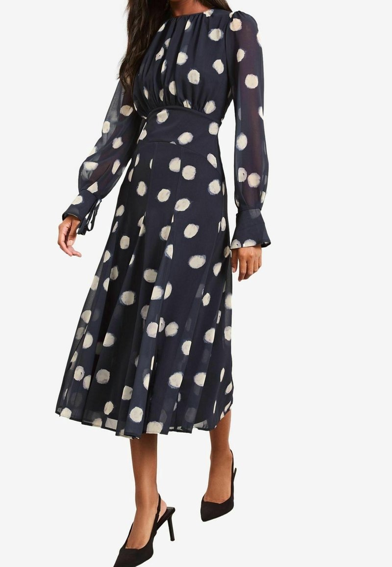 Robe midi bleu marine à pois blancs, manches longues en voile transparent, taille haute et jupe plissée, portée avec des talons noirs à bout pointu.