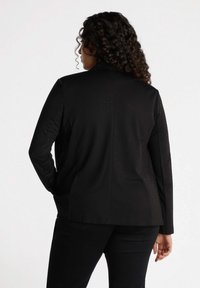 Svart blazer med prickig textur, enkel knappstängning, skräddarsydd passform och slagkrage. Tyget ser slätt ut med en lätt lyster.