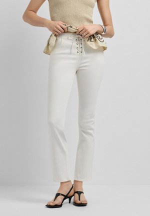 Donna che indossa pantaloni bianchi a vita alta con lacci, top senza maniche beige in maglia e sandali neri con tacco, che sistema il top in vita.