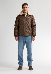 Hombre de pie mirando de frente, vistiendo una chaqueta de cuero marrón con cuello de borrego, jeans azul claro y botas de gamuza marrón, sobre un fondo blanco.