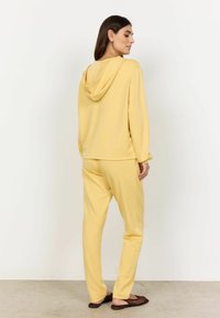 Geel lounge-outfit bestaande uit een hoodie en bijpassende broek, gemaakt van zachte stof met een gladde textuur, gedragen met bruine instap schoenen.