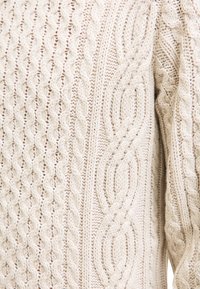 Ecrufarbenes Strickgewebe mit detailliertem Zopfmuster und strukturierten Motiven auf einem Nahaufnahme von einem Pullover.