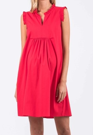 Mujer con vestido sin mangas de color rojo brillante con hombros con volantes y cintura fruncida, de pie contra un fondo claro y liso.