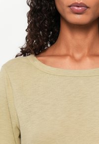 Lys olivengrønn topp med en svakt avrundet halslinning og mykt, teksturert stoff. Har et enkelt design uten mønstre eller dekorasjoner.