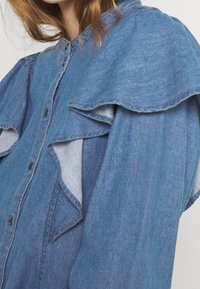 Jeanshemd mit Rüschendesign an den Schultern, in einer hellblauen Waschung und mit Knopfverschluss an der Vorderseite. Glatte Textur.