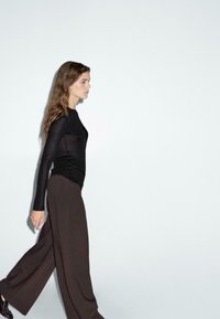 Zwarte langwerpige top met een aansluitend ontwerp, gecombineerd met brede, donkerbruine broek. Haar is golvend; het model loopt tegen een lichte achtergrond.