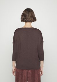 Personne aux cheveux courts et bruns portant une chemise ample à manches longues marron foncé et un pantalon marron texturé, vue de dos sur fond blanc.