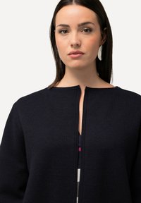 Mörkblå stickad cardigan med rund halsringning och rak passform. Har en central öppning och texturerad kvalitet. Bärs över en mönstrad skjorta.