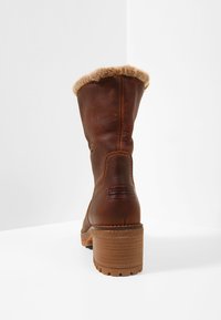 Bota de cuero marrón con puño de pelo sintético, tacón cuadrado, y logo embossado en la parte posterior. Textura suave con detalles de costura a lo largo de los lados.