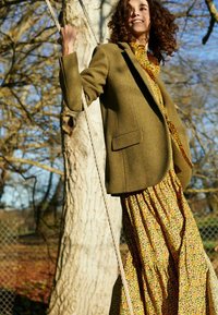 Blazer vert olive sur une robe maxi jaune à fleurs avec volants. Tissu doux, silhouette structurée et motif floral élaboré. Arrière-plan naturel.
