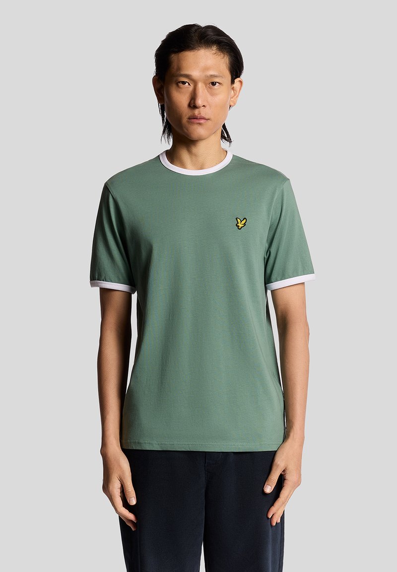 Un uomo sta in piedi di fronte, indossando una maglietta verde salvia con bordo bianco e un piccolo logo di un uccellino giallo sul petto, pantaloni scuri.
