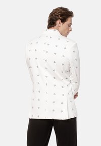 Mężczyzna w białym żakiecie z czarnym abstrakcyjnym wzorem, stojący z założonymi rękami, zwrócony plecami, widoczne czarne spodnie.