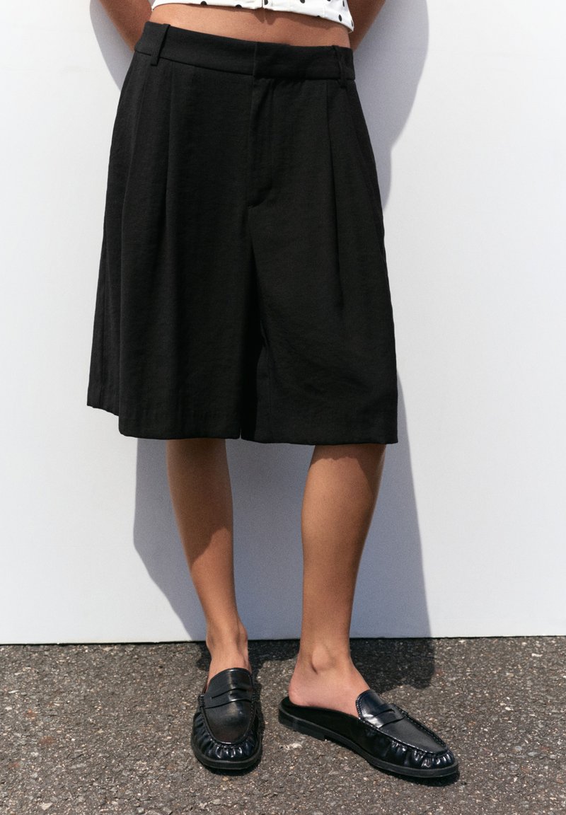 Partie inférieure du corps d'une personne portant un short noir ample jusqu'aux genoux et des mocassins noirs sans lacets, debout sur de l'asphalte sur fond blanc.