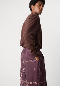 Blusa de manga larga marrón combinada con pantalones morados oscuros que presentan un estampado de un mapa de Lagos, con bolsillos y un cinturón como acento. Textura suave.