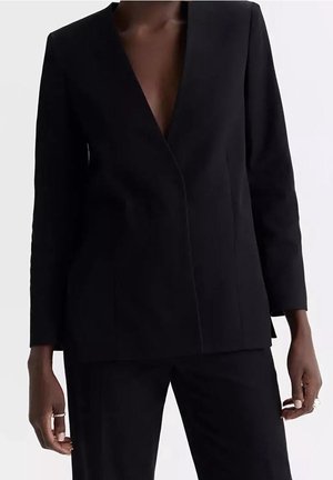 Kvinde model iført et skræddersyet sort jakkesæt med en dyb V-hals blazer og matchende sorte bukser, stående mod en hvid baggrund.