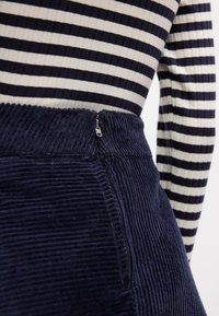 Gros plan sur une personne portant une jupe en velours côtelé bleu marine avec une fermeture éclair latérale et un haut à manches longues à rayures horizontales noires et blanches.
